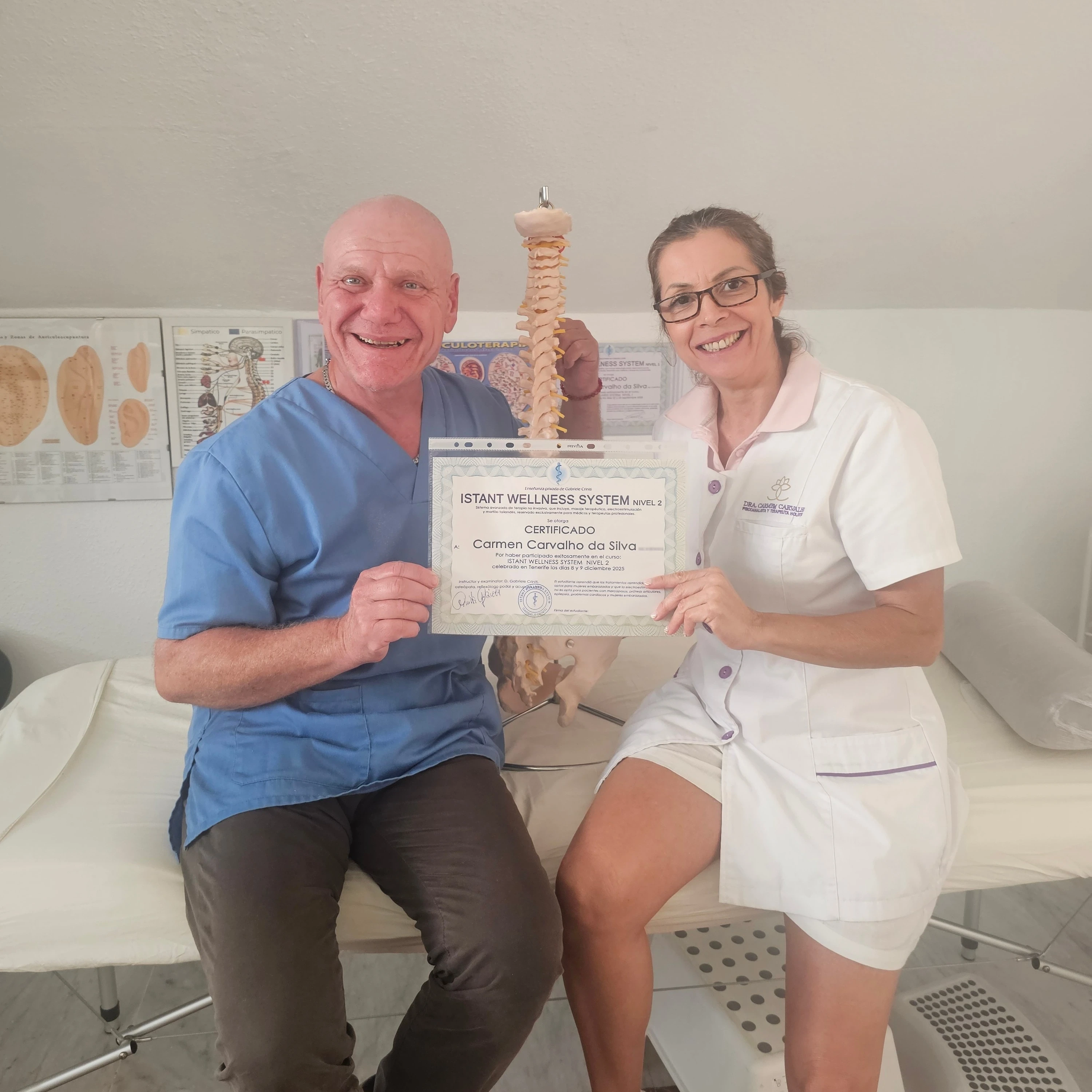 Carmem Carvalho con uniforme sanitario blanco recibiendo el certificado de Acupuntura Clínica y Terapia Manual junto a su formador, en la camilla del centro de formación