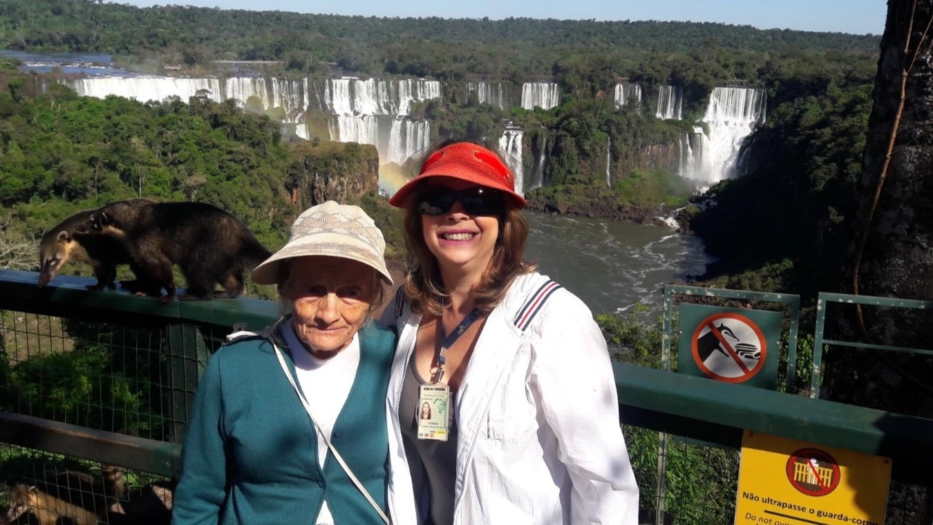 Carmem Carvalho durante sus años como guía turística internacional, junto a una mujer mayor frente a las cataratas de Iguazú — la vida que parecía libertad antes de aprender a escuchar al cuerpo